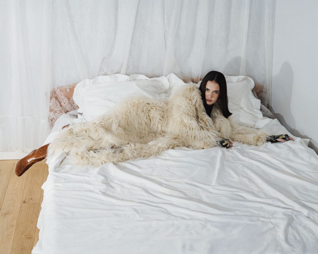 Młoda kobieta o ciemnych włosach leży na łóżku z białą pościelą. Ma na sobie obszerny, biały, futrzany lub wełniany płaszcz o długim włosiu. Kobieta patrzy bezpośrednio w obiektyw aparatu, opierając się na łokciu.
